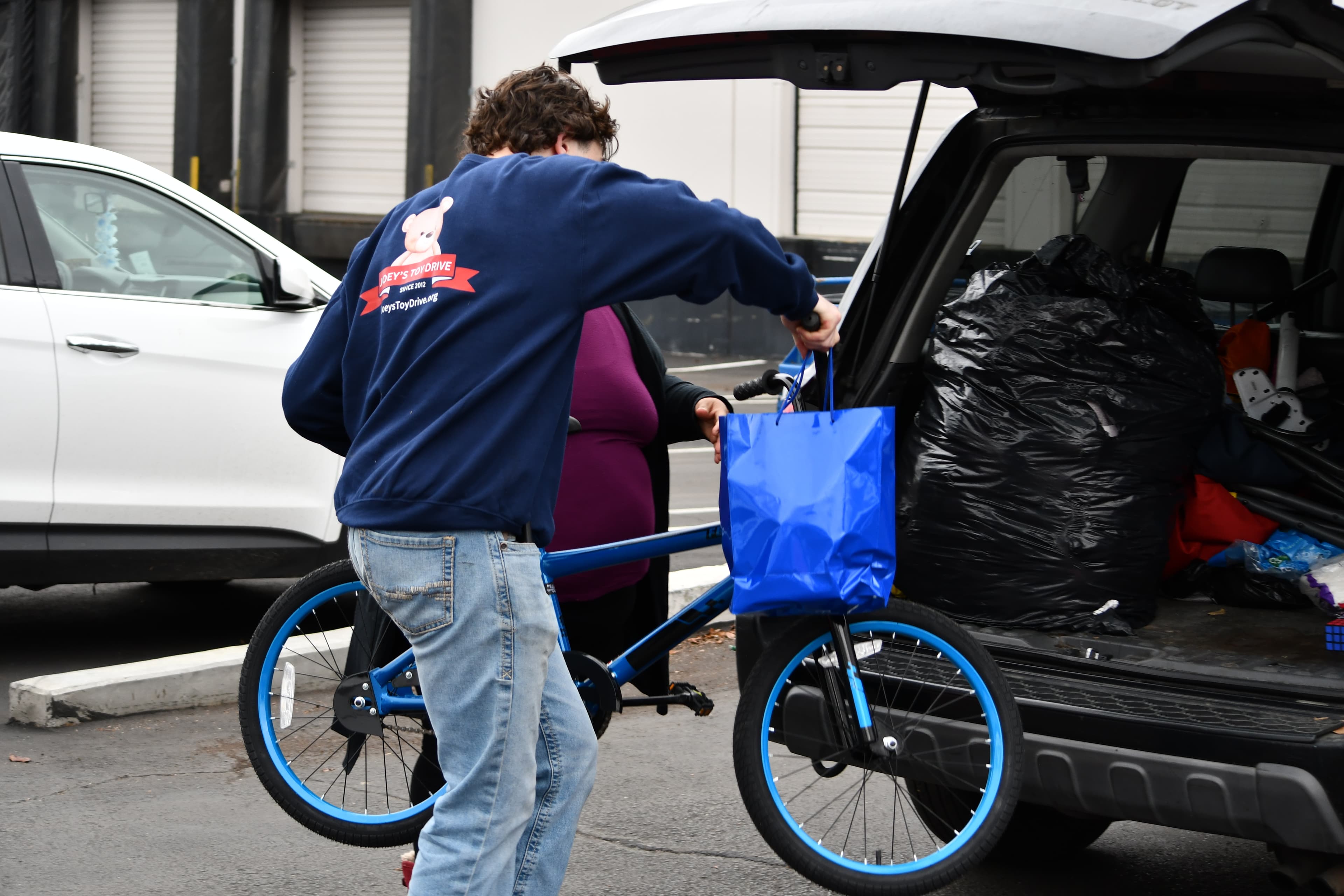 A volunteer in a Joey's Toy Drive sweatshirt loading a child's bicycle and a JTD gift bag into the back of a car for delivery