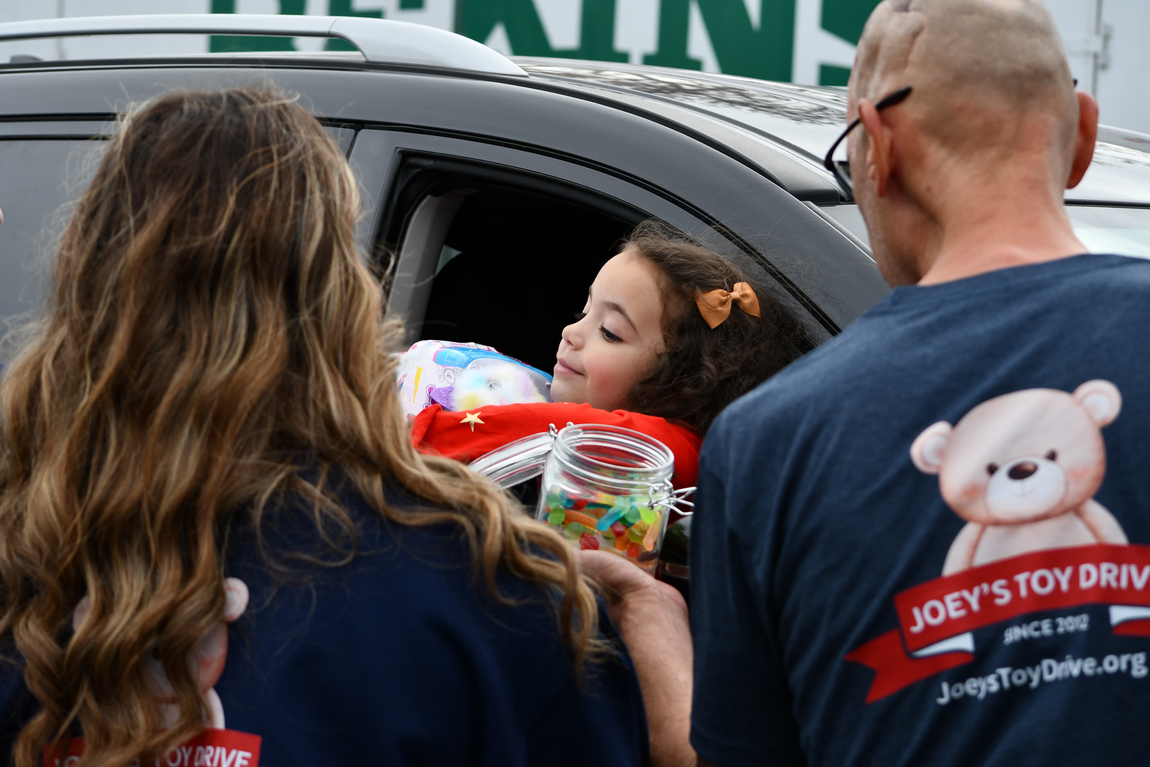 A child in the passenger seat of a car receives a jar of toys from two Joey's Toy Drive volunteers — the bear logo and wordmark visible on the back of one volunteer's shirt
