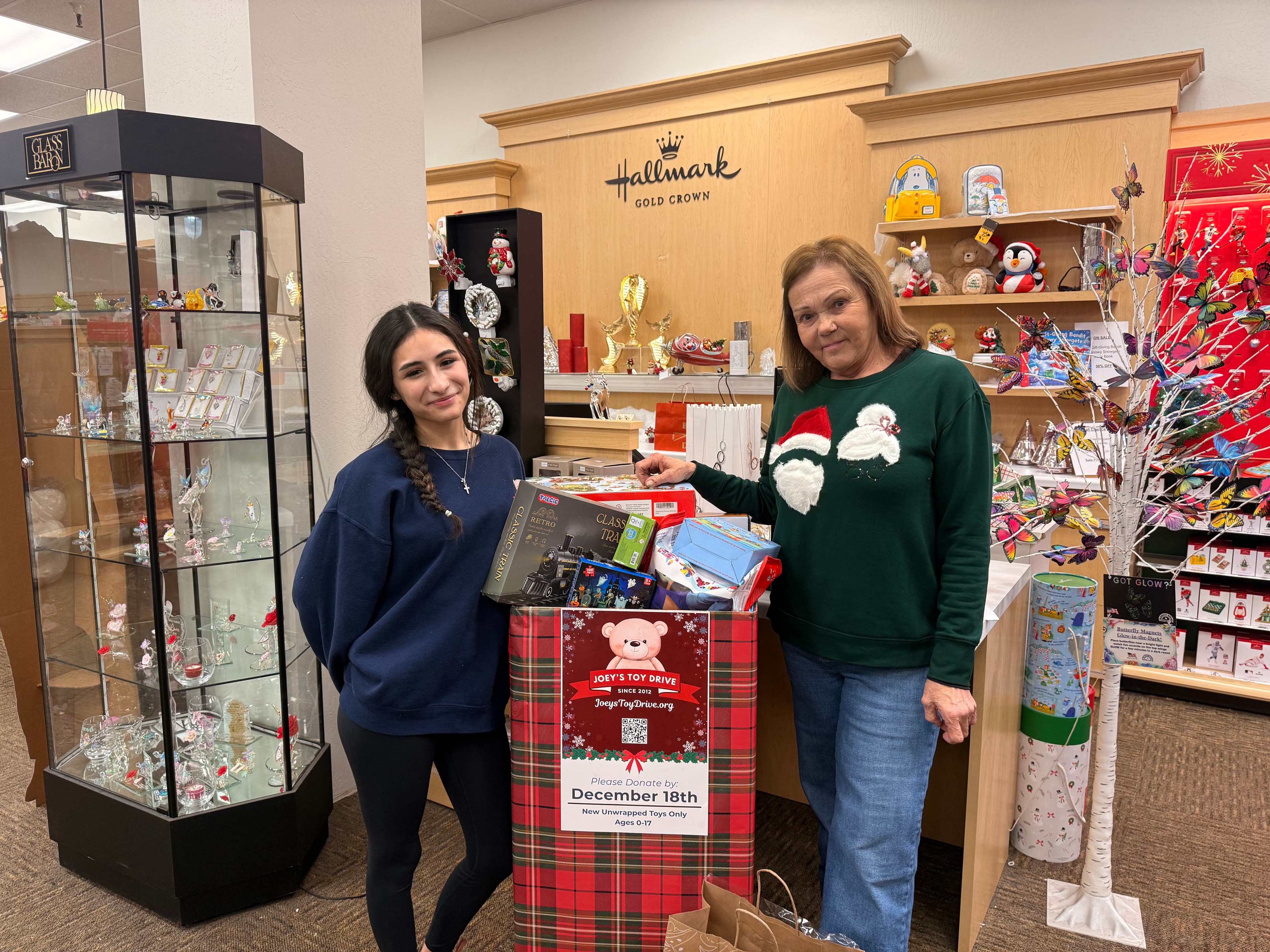 Two volunteers standing beside a Joey's Toy Drive donation box at a partner storefront, with toys piling up inside the branded container