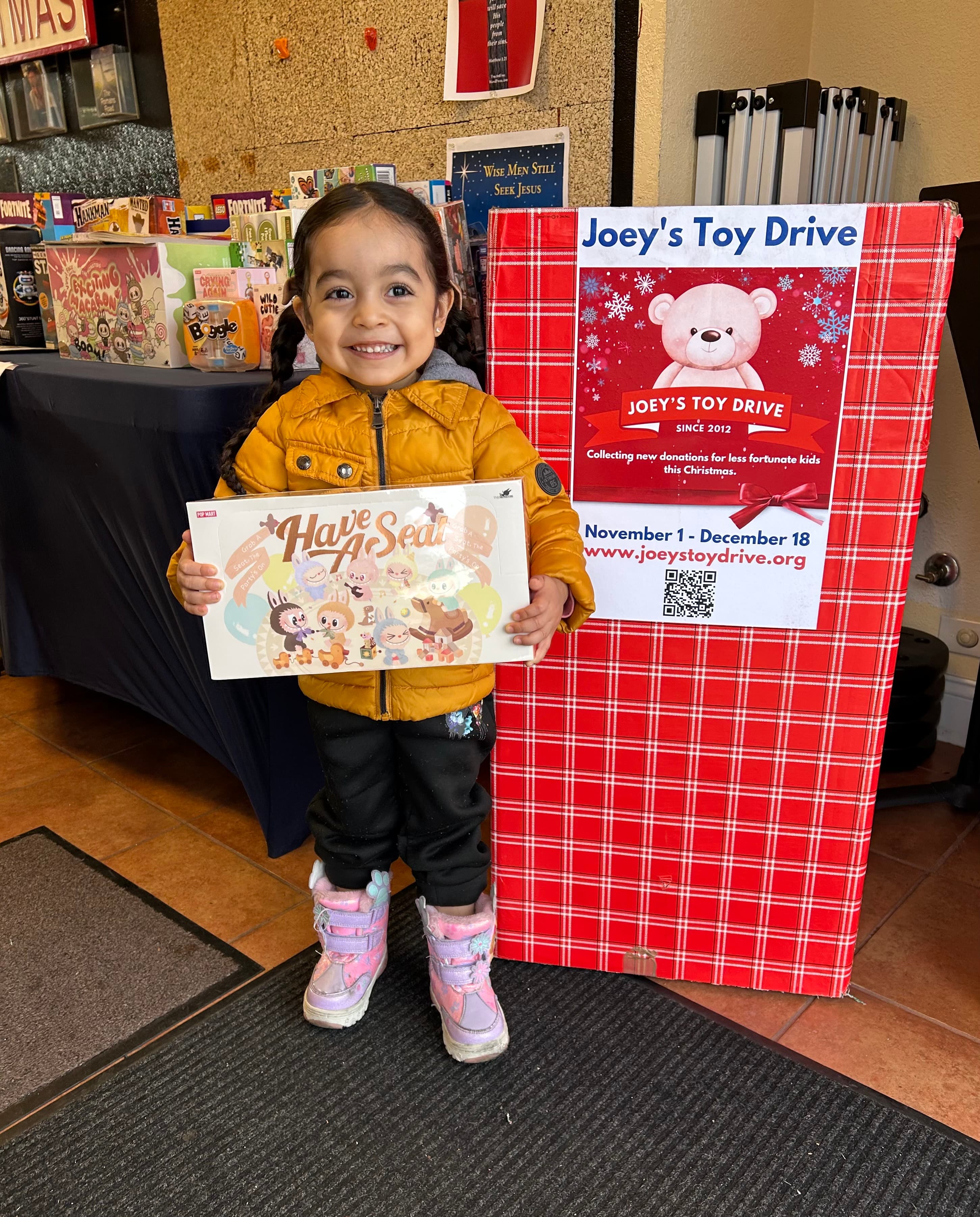 A young girl smiling beside a Joey's Toy Drive donation box, holding a 'Have A Seat' picture book she just received