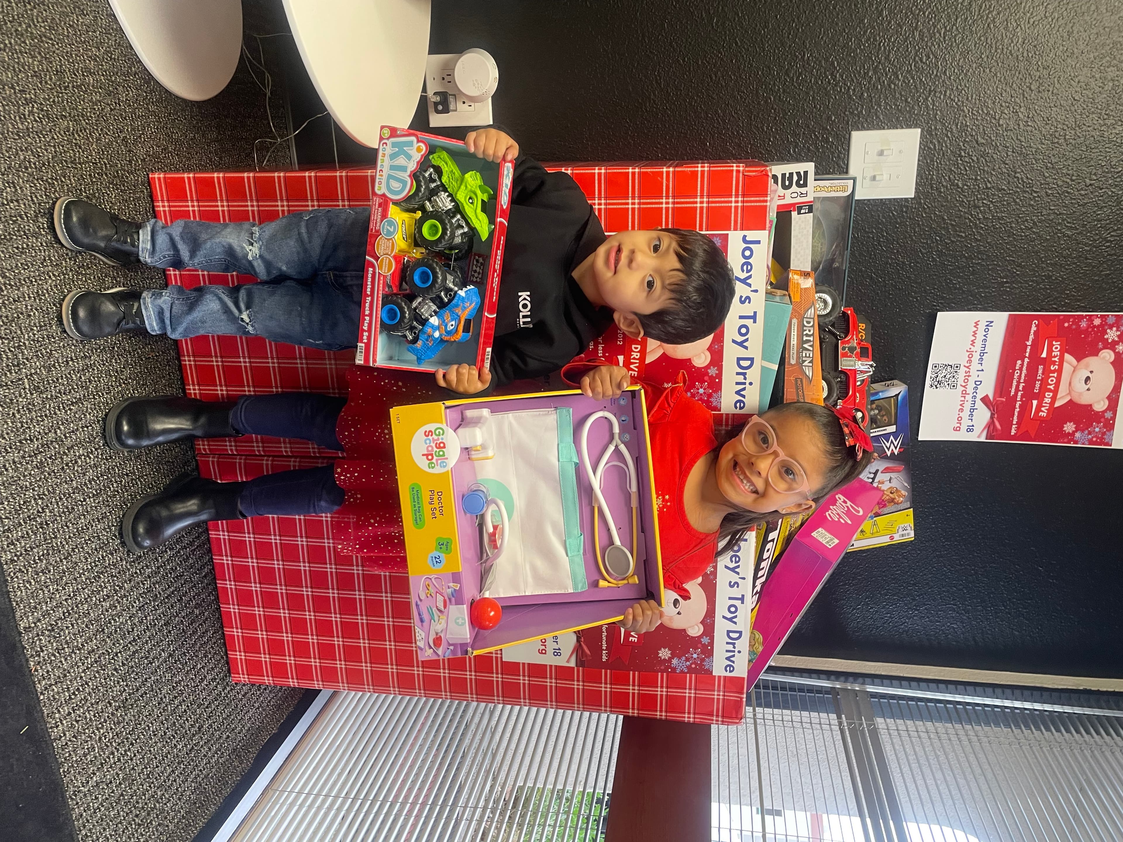 Two children holding the new toys they just received next to a Joey's Toy Drive donation box